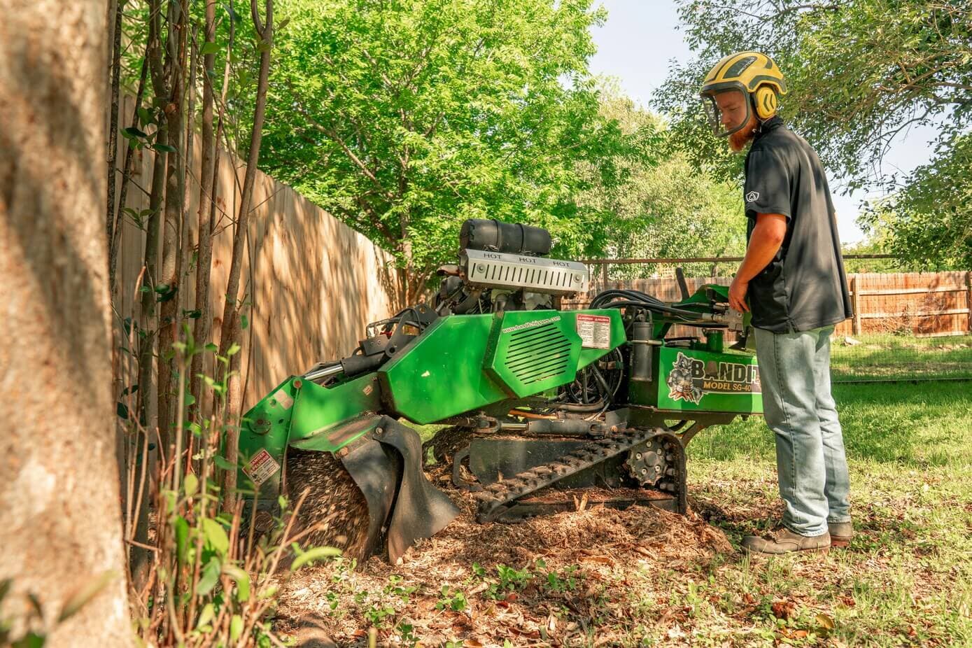 Worker operating a compact stump grinder beside a wooden fence
